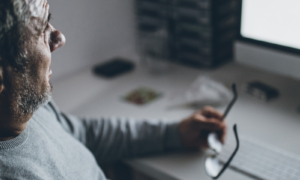 Worried senior man sitting in front computer, holding his glasses