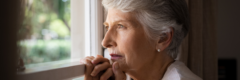 Depressed senior woman at home feeling sad. Elderly woman looks sadly outside the window. Depressed lonely lady standing alone and looking through the window.