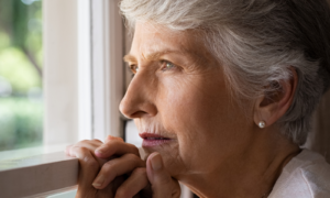 Depressed senior woman at home feeling sad. Elderly woman looks sadly outside the window. Depressed lonely lady standing alone and looking through the window.