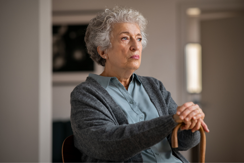 Retired unhappy woman with her wooden walking stick at home. Lonely serious senior woman holding walking cane and looking through the window. Moody and upset grandmother sitting on couch in nursing home.