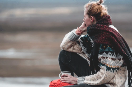 Woman in a jumper and scarf, with a bun in her hair, reflecting on life and death, while drinking a coffee.